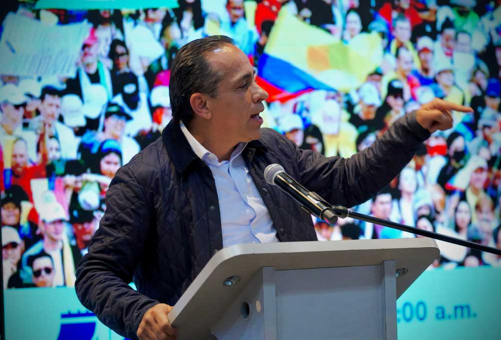 Director del DNP, Alexander López en tarima dando un discurso. De fondo una pantalla gente con la bandera de Colombia.